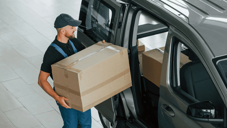 This image shows a delivery worker lifting a large cardboard box from the back of a truck. The worker is wearing a navy blue uniform, a cap, and glasses, while focusing on the task of moving the box. The background includes other similar boxes, indicating a busy loading or unloading process. The worker appears to be handling the task with care, ensuring that the package is moved safely.