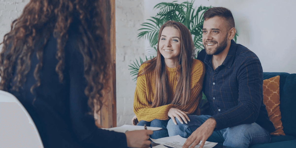 A couple smiles while listening to a person seated across from them in a cozy setting