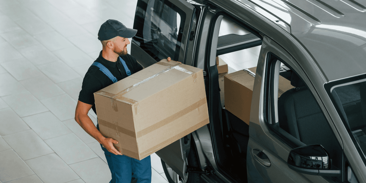 This image shows a delivery worker lifting a large cardboard box from the back of a truck. The worker is wearing a navy blue uniform, a cap, and glasses, while focusing on the task of moving the box. The background includes other similar boxes, indicating a busy loading or unloading process. The worker appears to be handling the task with care, ensuring that the package is moved safely.