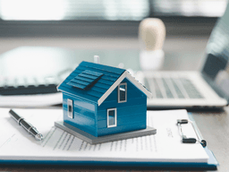 A blue model house on paperwork with a laptop and calculator in the background