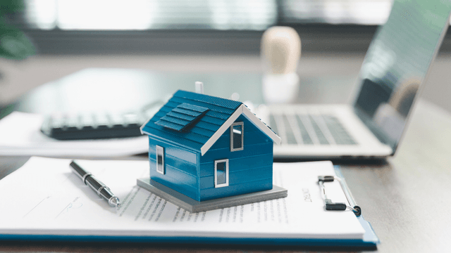 A blue model house on paperwork with a laptop and calculator in the background