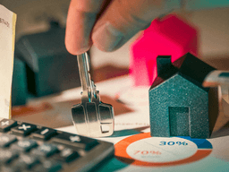 A person holding keys over miniature houses and financial charts with currency and calculator