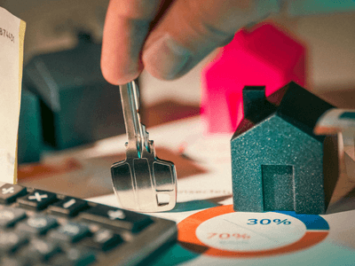 A person holding keys over miniature houses and financial charts with currency and calculator