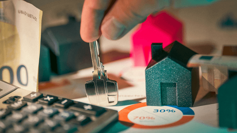 A person holding keys over miniature houses and financial charts with currency and calculator