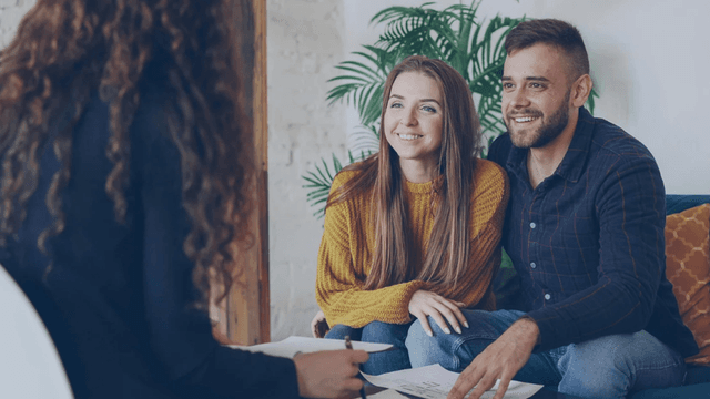 A couple smiles while listening to a person seated across from them in a cozy setting