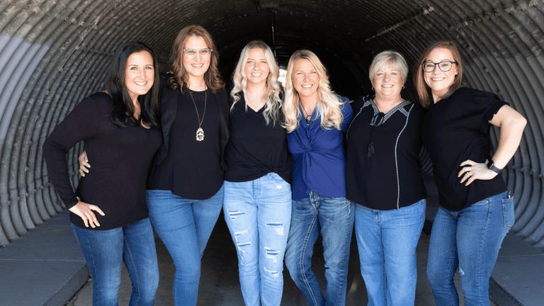 "A group portrait of six women standing together in a tunnel-like space. They are dressed in black tops and denim jeans, smiling confidently and showing camaraderie. The photo captures their strong bond and positive energy."