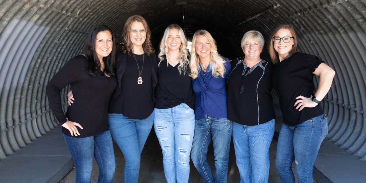 "A group portrait of six women standing together in a tunnel-like space. They are dressed in black tops and denim jeans, smiling confidently and showing camaraderie. The photo captures their strong bond and positive energy."