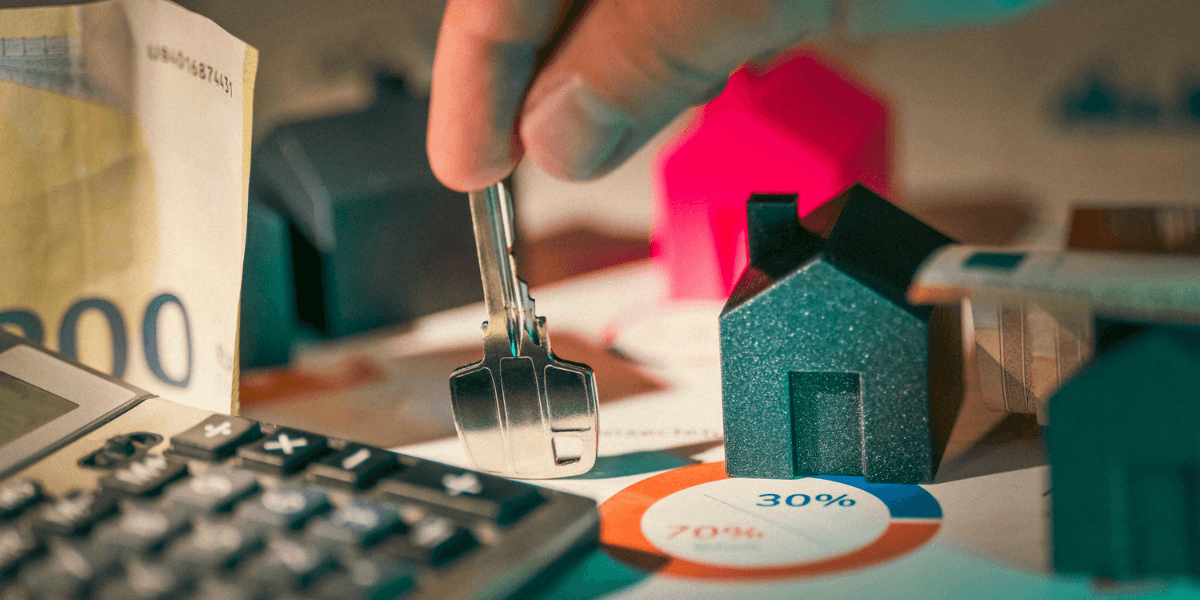 A person holding keys over miniature houses and financial charts with currency and calculator